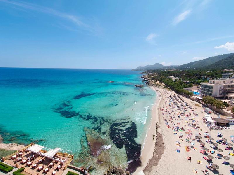 Beach with clear blue sea, sunbathers, and hotels along the shore under a bright sky.