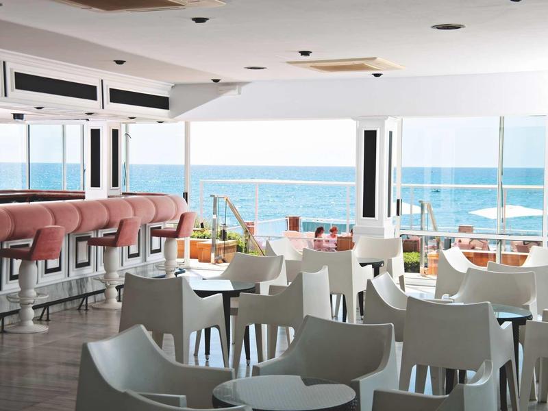 Modern beachfront café with white chairs, rounded tables, and pink bar stools with ocean view.