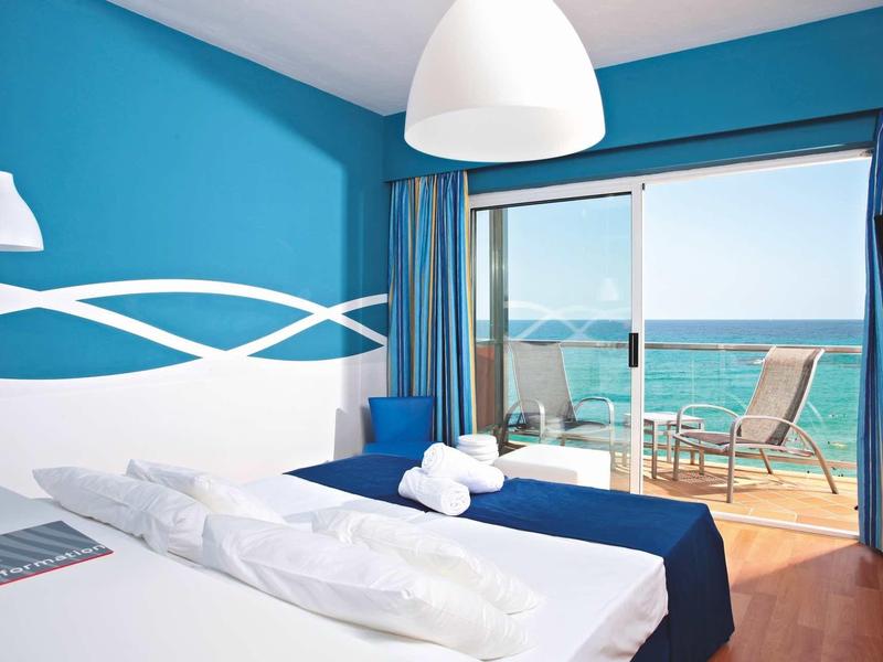 Bright hotel room with blue walls, white bed linens, and balcony overlooking the sea.
