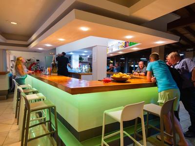 Bar moderna in hotel con bancone illuminato e sedie, ospiti seduti e in piedi.