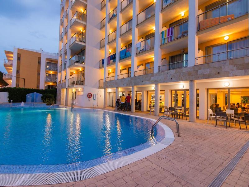 Piscina esterna illuminata davanti a un edificio alberghiero a più piani al crepuscolo.