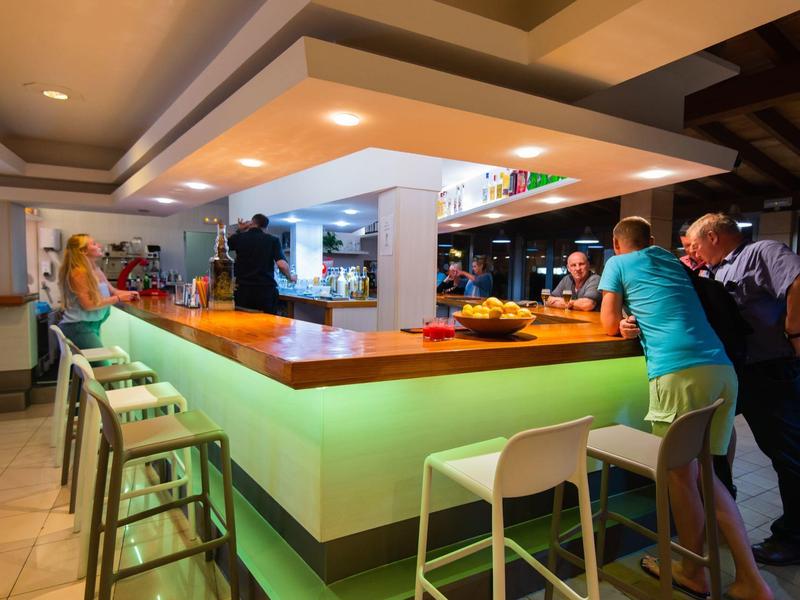 Bar moderna in hotel con bancone illuminato e sedie, ospiti seduti e in piedi.