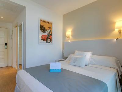 Hell eingerichtetes Schlafzimmer mit Doppelbett, weißen Wänden, grauer Bettdecke und Wandlampen.