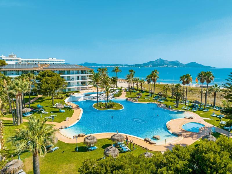 Duży basen hotelowy z palmami i widokiem na morze i góry w tle.