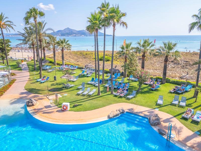 Basen hotelowy z leżakami i palmami obok piaszczystej plaży i morza w tle.