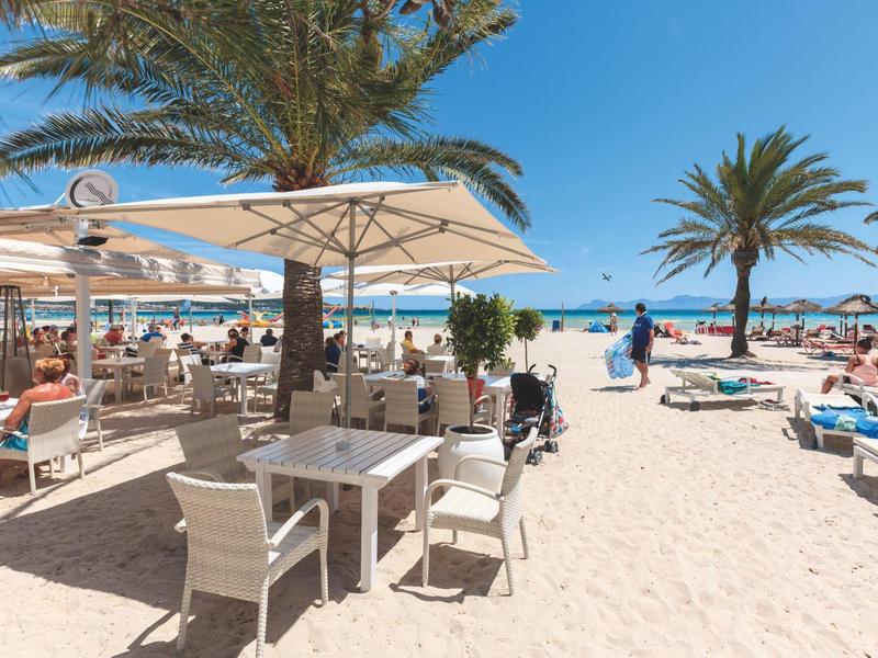 Strefa do posiłków na plaży z stołami, krzesłami, parasolami, palmami i leżakami na piasku.