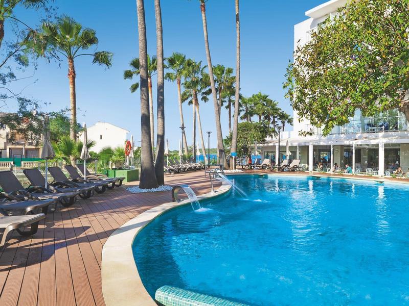 Basen hotelowy na zewnątrz otoczony leżakami i wysokimi palmami w słoneczny dzień.