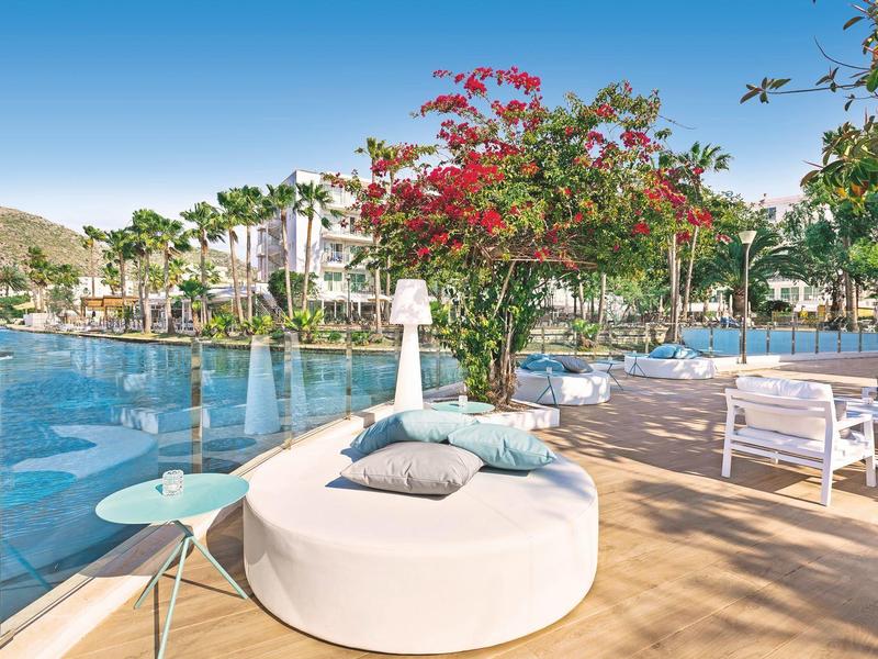 Słoneczny teren przy basenie na zewnątrz z okrągłym leżakiem, szklaną barierką i kolorowymi drzewami.