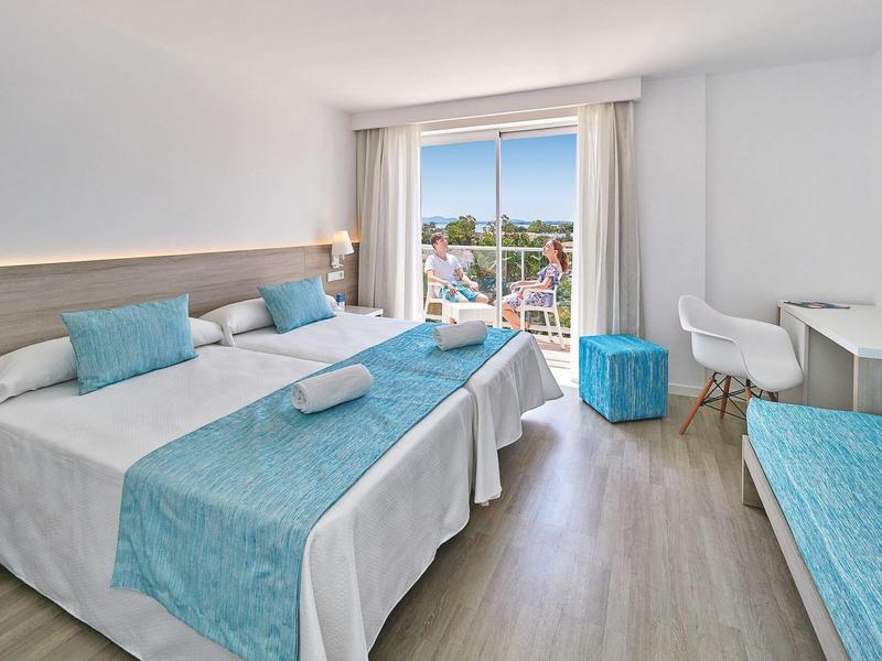 Jasny pokój hotelowy z dwoma łóżkami, niebieskimi dodatkami, balkonem i nowoczesnym wyposażeniem.