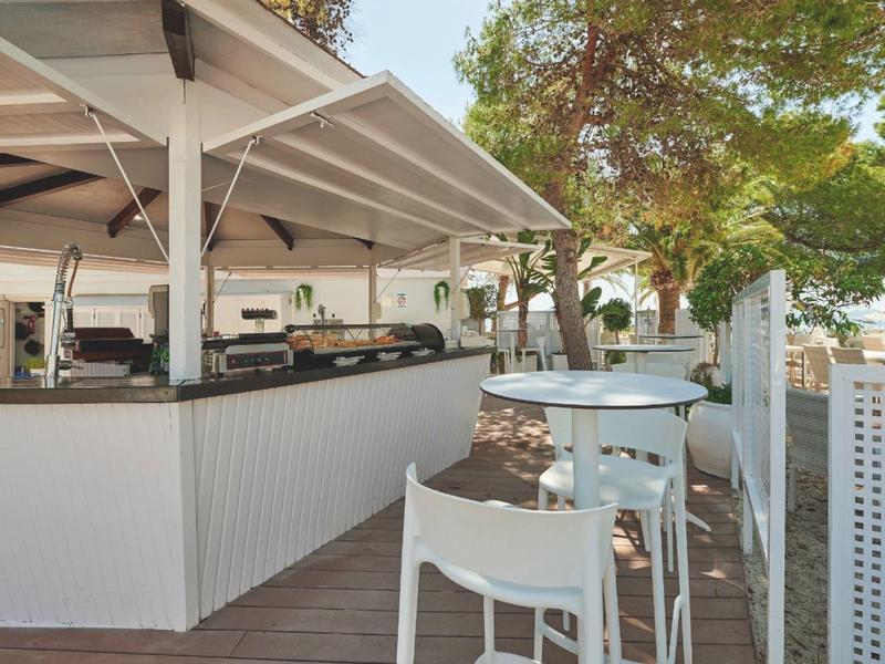 Zewnętrzny bar z białymi wysokimi krzesłami i stolikami pod markizą obok drzew i drewnianej podłogi.