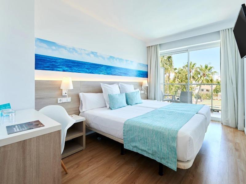 Jasny pokój hotelowy z podwójnym łóżkiem, niebieskim wystrojem, drewnianą podłogą i balkonem z widokiem na morze.