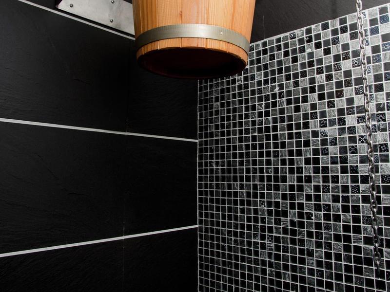 Holzeimer-Dusche an schwarzer Wand mit grauen Mosaikfliesen und Chromkettenhalterung.