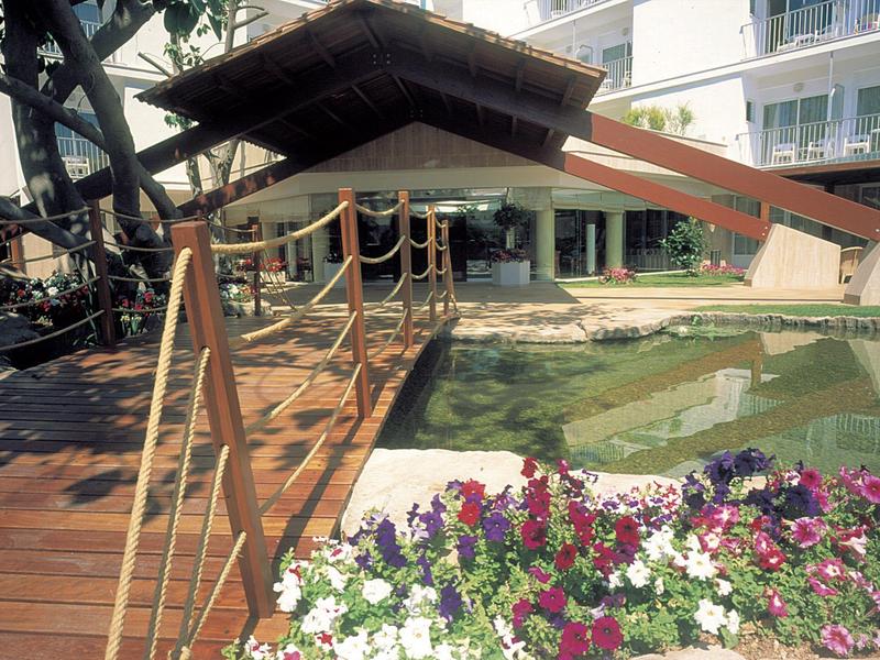 Holzbrücke mit Tau-Geländer über Teich, daneben bunte Blumen und modernes Gebäude im Hintergrund.