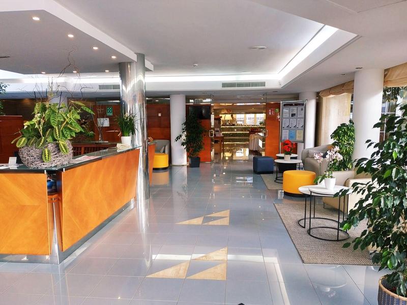 Moderne, helle Empfangshalle mit grauem Boden, gelben Sitzmöbeln und vielen grünen Pflanzen.