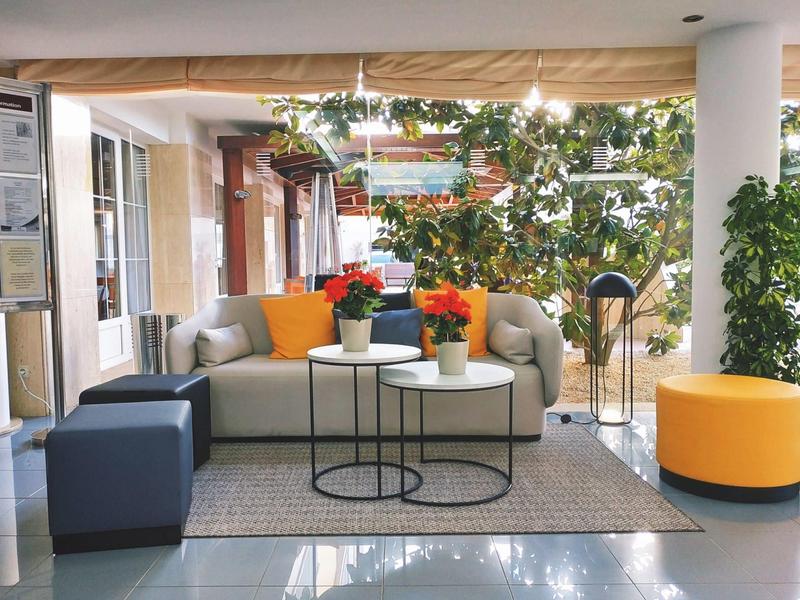 Moderner Sitzbereich mit grauer Couch, bunten Kissen, Pflanzentöpfen und Teppich auf glänzendem Boden.