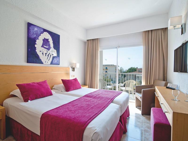 Hell eingerichtetes Hotelzimmer mit Doppelbett, lila Akzenten und Balkon mit Meerblick.