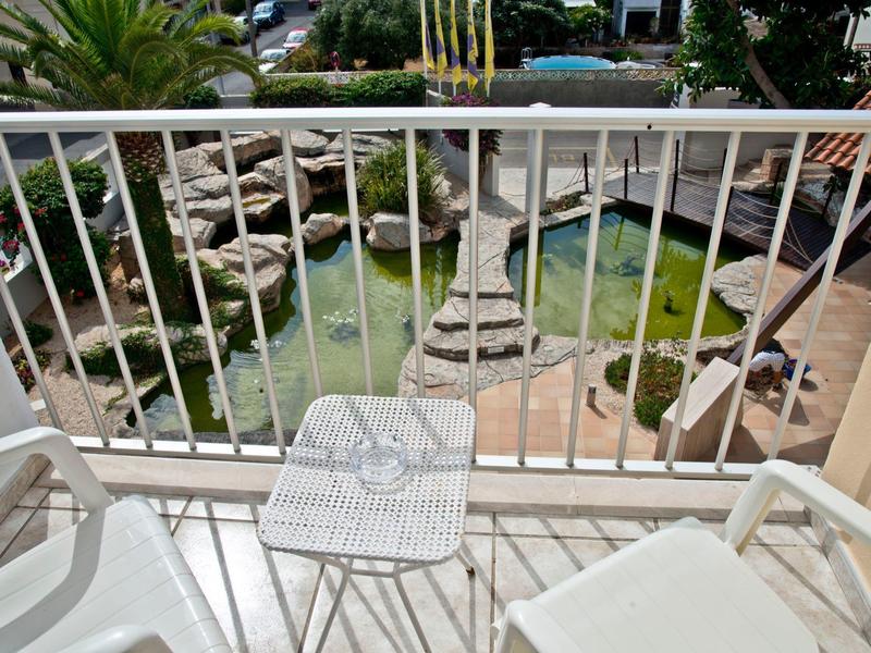 Balkon mit weißen Stühlen und Tisch, Blick auf Teich mit Steinen und grünen Palmen im Hintergrund.