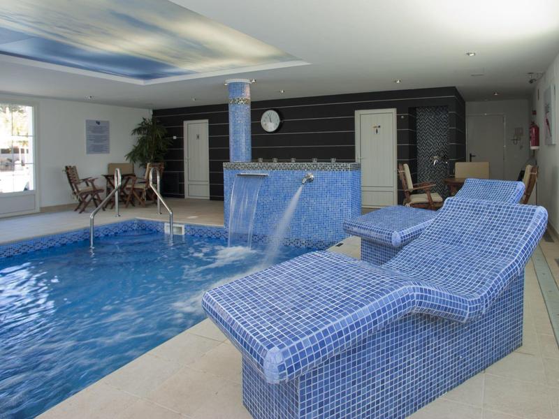 Innenpool mit blauen Mosaik-Liegen, Wasserspielen und großen Fenstern, helle Atmosphäre.