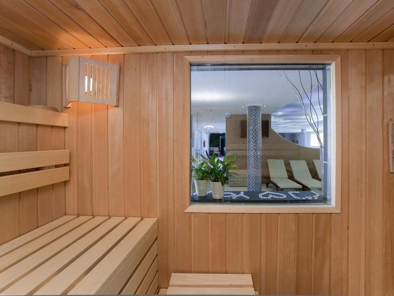 Sauna mit hölzernen Sitzbänken, Wandlampen und Fenster mit Blick in einen Raum mit Pflanzen.