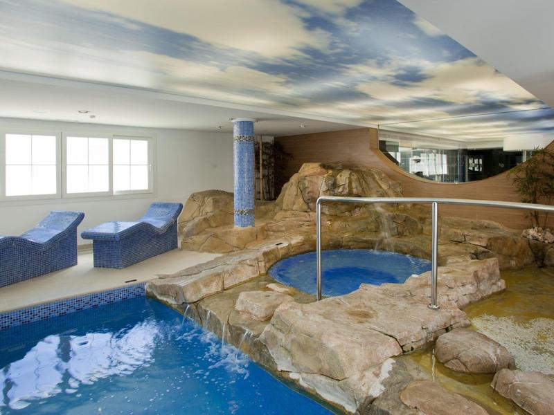 Innenraum mit Pool, Felsenimitat, blauen Liegen und Himmel-Decke mit Wolkenmotiv.