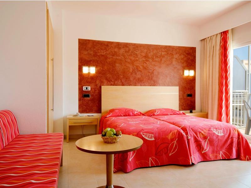 Helles Schlafzimmer mit roten Bettdecken, roten Vorhängen, Sofa und Balkon mit Meerblick.