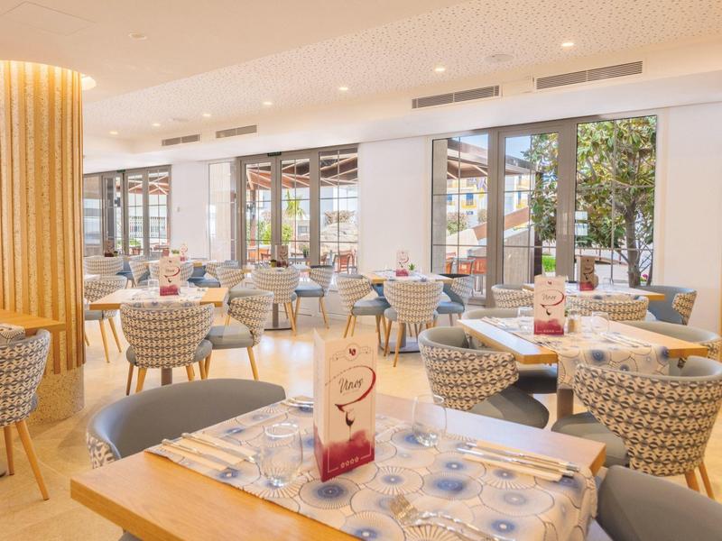 Hell eingerichtetes Restaurant mit runden Tischen, gepolsterten Stühlen und großen Fenstern.