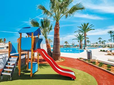 Barevné hřiště u bazénu s palmami pod jasně modrou oblohou.