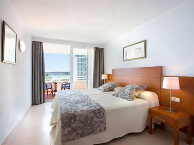 Světly hotelový pokoj s manželskou postelí, nočními stolky a balkonem s výhledem na moře.