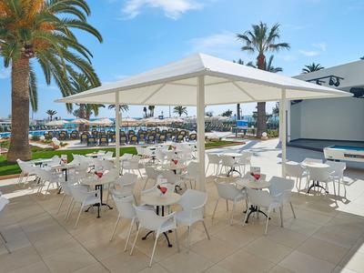 Venkovní restaurace s bílými stoly a židlemi vedle bazénu a palem.