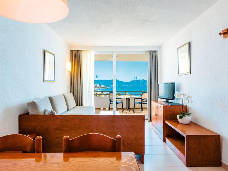 Světý obývací pokoj s pohovkou, TV a výhledem na moře přes balkonové dveře.