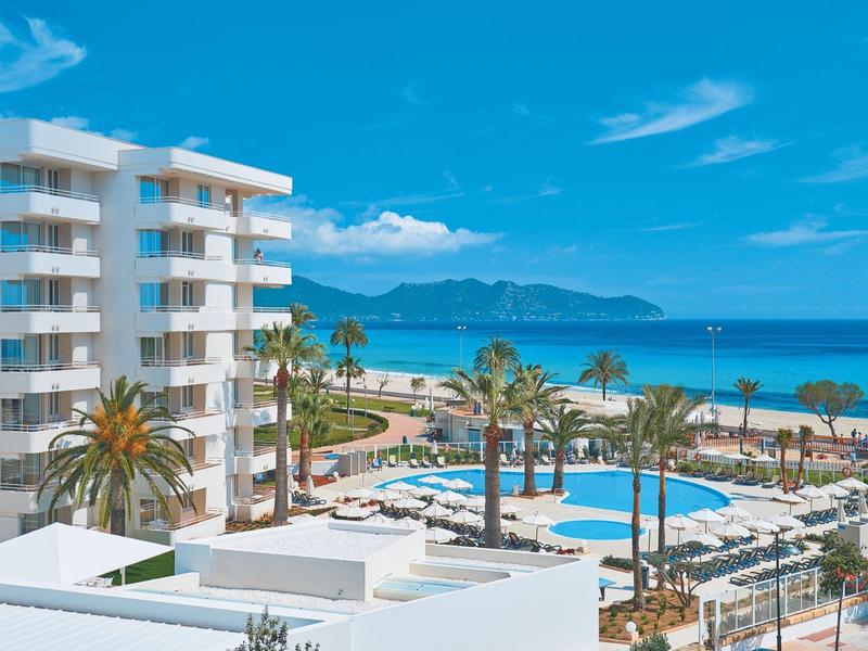 Bílý hotel s bazénem, palmami a výhledem na moře pod světlou modrou oblohou.