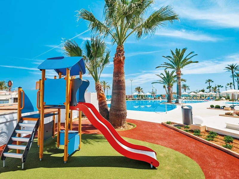 Barevné hřiště u bazénu s palmami pod jasně modrou oblohou.