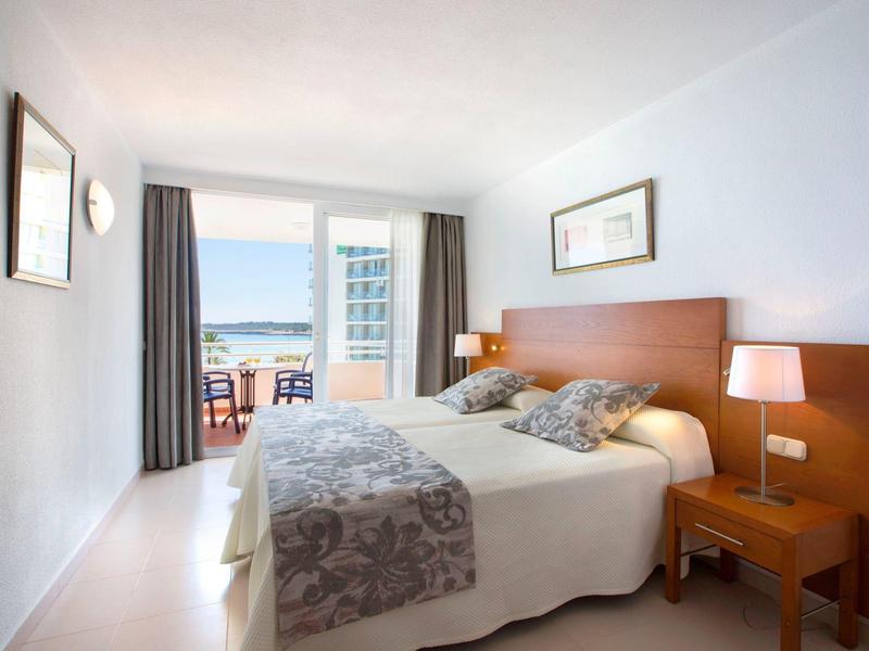 Světly hotelový pokoj s manželskou postelí, nočními stolky a balkonem s výhledem na moře.