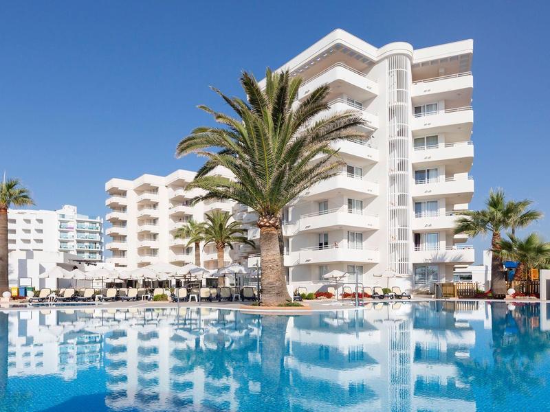 Hotel s bílými budovami a velkým bazénem pod jasnou modrou oblohou.