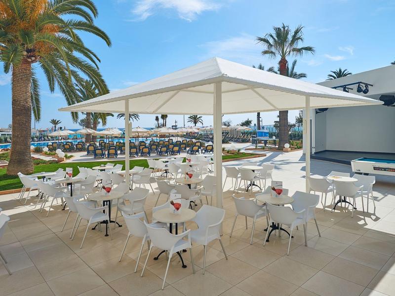 Venkovní restaurace s bílými stoly a židlemi vedle bazénu a palem.