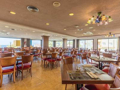 Ruime, lichte eetzaal met houten tafels en stoelen en grote ramen met uitzicht.