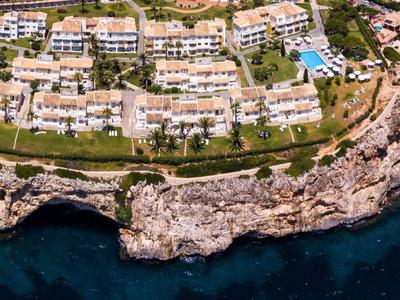 Kustresort met witte gebouwen, groene gazons, zwembaden en rotsachtige kustlijn bij diepblauwe zee.