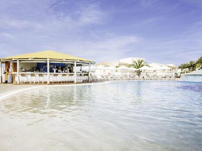 Groot zwembad met paviljoen en palmbomen onder een blauwe lucht in een vakantieoord.