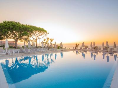 Infinity-zwembad met uitzicht op de stadsgevel bij zonsondergang, omringd door ligstoelen en bomen.