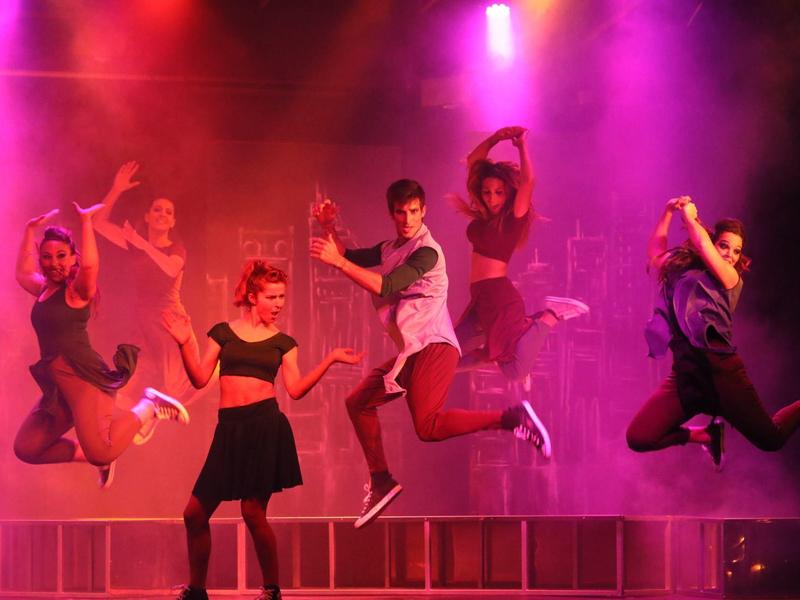 Vijf mensen dansen energiek op een podium met kleurrijk licht op de achtergrond.