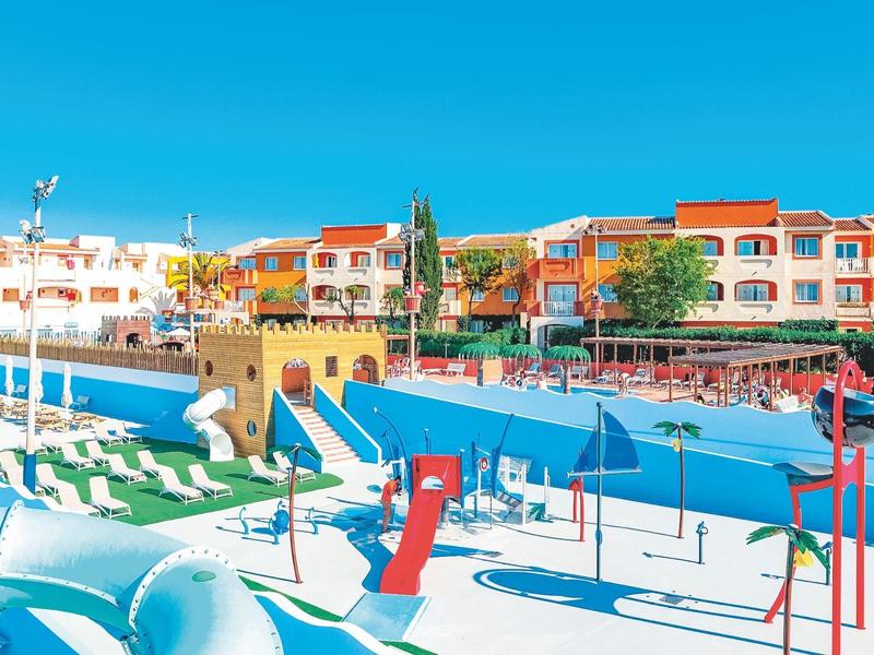 Kleurrijk hotel met zwembadgebied en parasols onder een heldere blauwe lucht.