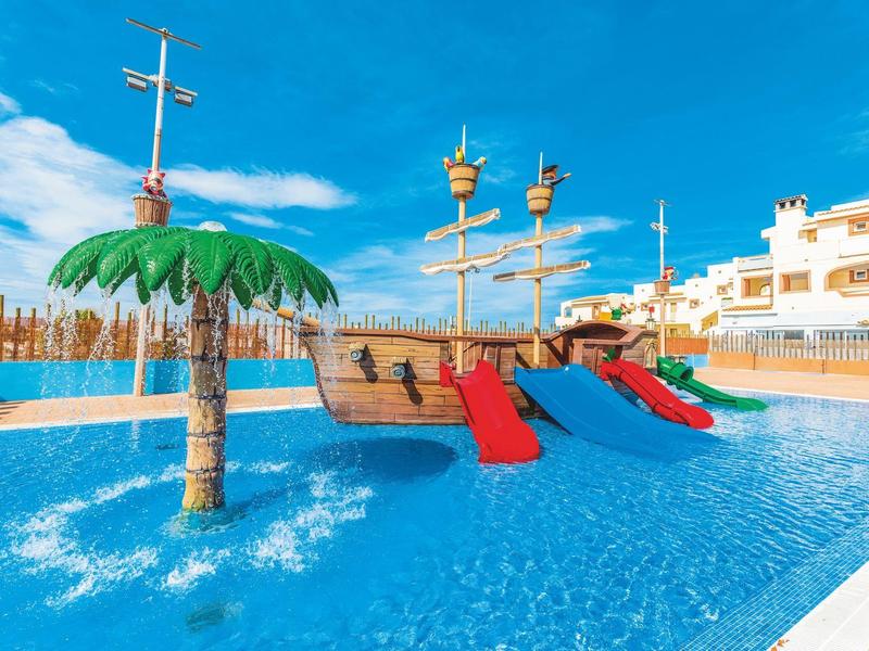 Kinderzwembad met piratenschipglijbanen en palmbomen onder een zonnige hemel bij een resort.