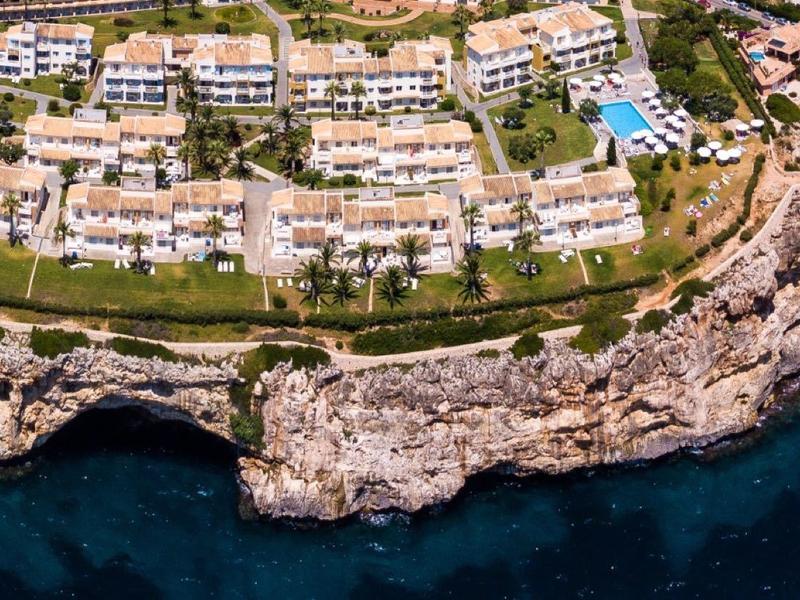 Kustresort met witte gebouwen, groene gazons, zwembaden en rotsachtige kustlijn bij diepblauwe zee.