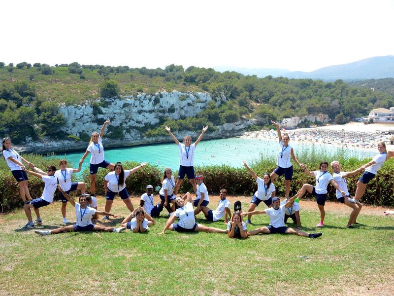 Een groep mensen doet acrobatiek op gras bij water met bomen op de achtergrond.