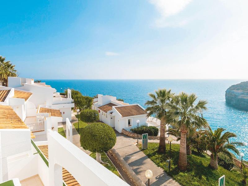 Witte huizen met terrassen aan zee onder een heldere blauwe lucht met palmbomen en kliffen.