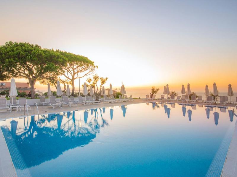 Infinity-zwembad met uitzicht op de stadsgevel bij zonsondergang, omringd door ligstoelen en bomen.