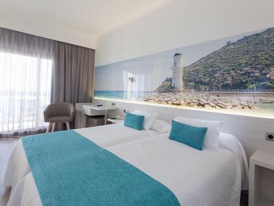 Nowoczesny pokój hotelowy z podwójnym łóżkiem, turkusowymi poduszkami i muralem latarni morskiej.