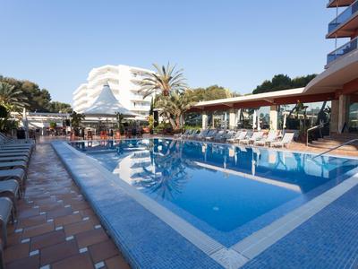 Strefa na zewnątrz basenu z leżakami, parasolami i wielopiętrowym hotelem w tle.