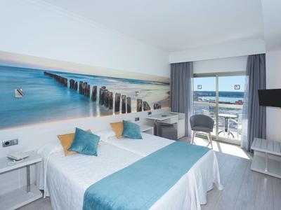 Camera d'hotel luminosa con due letti singoli, decorazione blu-bianca e balcone con vista mare.