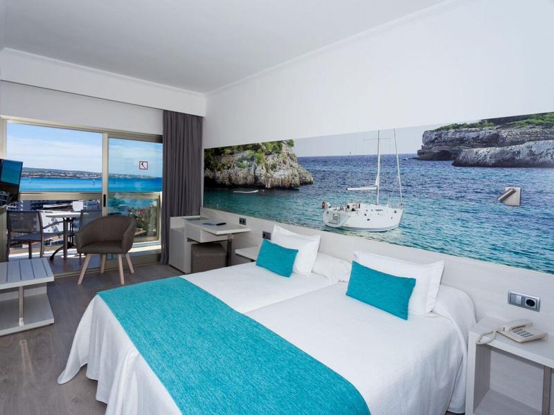 Nowoczesny pokój hotelowy z podwójnym łóżkiem i dużym muralem ściennym przedstawiającym zatokę morską z żaglówkami.